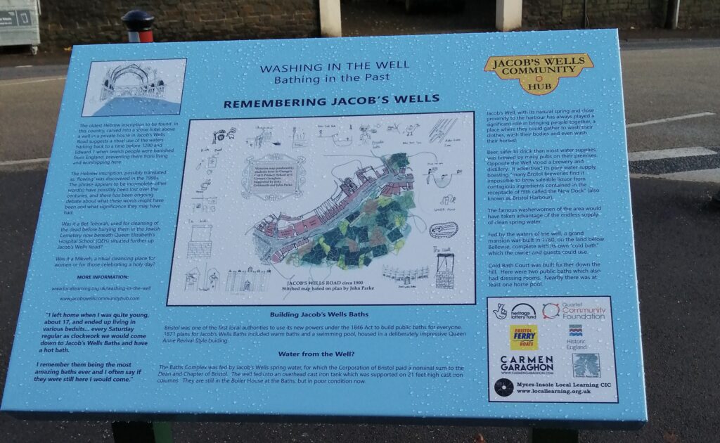 Information board standing in the street, titled “Washing in the Well -Bathing in the Past - Remembering Jacob’s Wells”
