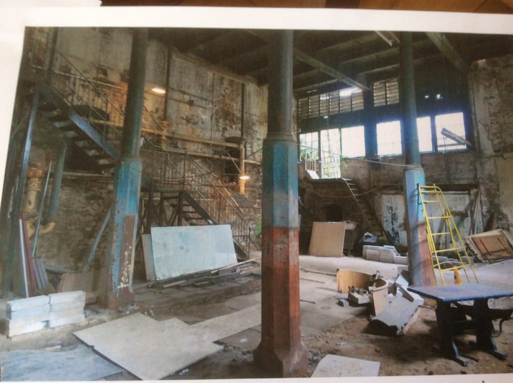 Photo of the boiler room, showing a double height room, brick walls, 4 largepillars, a steel staircase coming down, and a lot of rubble