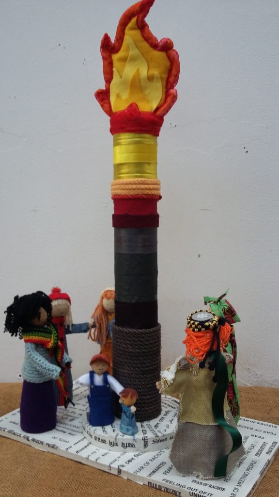 Photo of a
craft-piece made from fabrics over wood: a fiery beacon is aloft, with 5 adults and 2 children around the
base, from different backgrounds, on a base covered in phrases in different languages.