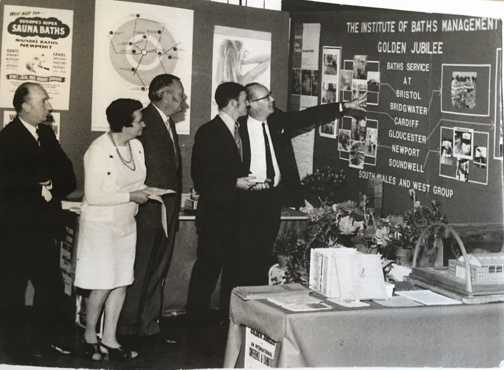 Smartly dressed men point at board with bath posters and text “The Instituteof Baths Management golden jubilee”